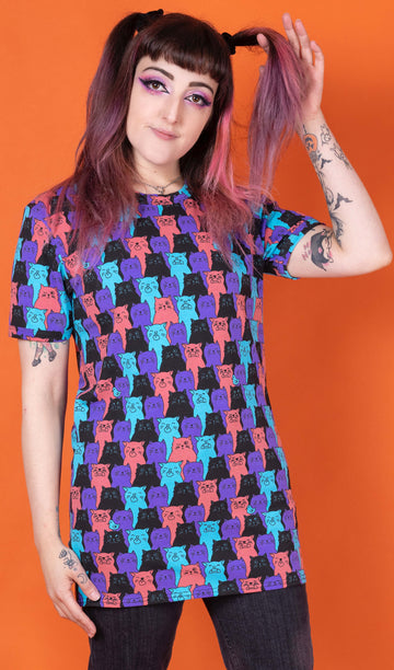 Sarah is stood in front of an orange background wearing the Cat Chorus Short Sleeve Tee with black stonewash denim flares. She is facing forward with one hand flicking her hair and the other hand resting by her side. The cat chorus print is an all over repeating pattern of sky blue, peachy red, purple and black cats pulling various faces and a sporadic sky blue bird.