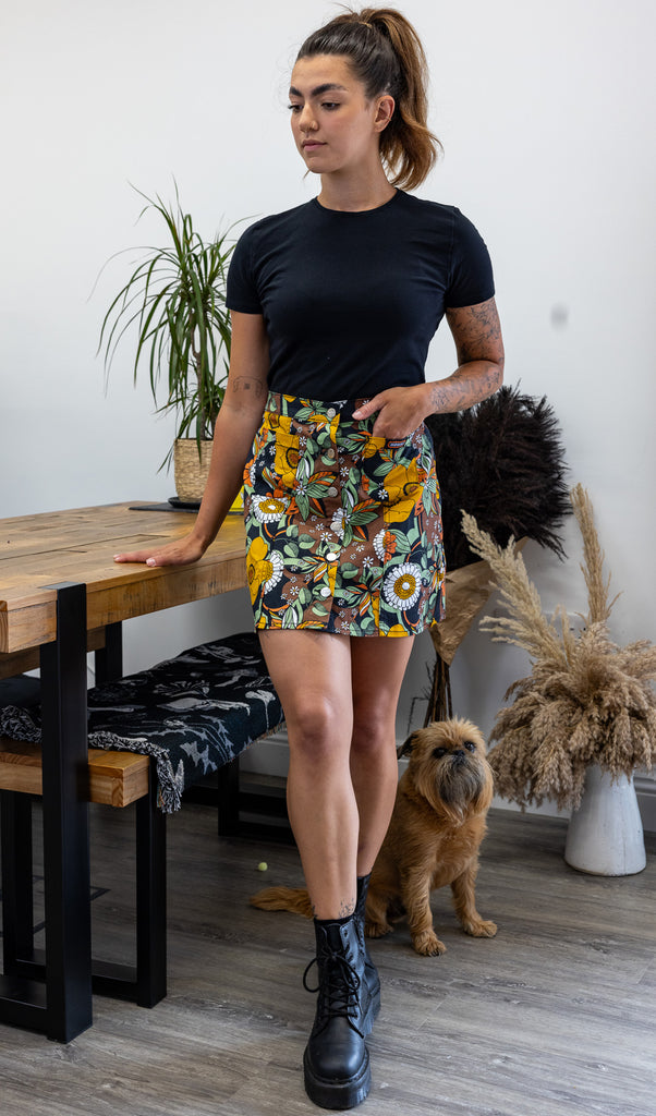 Model wearing 70's Black Floral A Line Skirt with a black t-shirt and boots. Model has her hand in the front pocket of the skirt. The skirt has a brown background with various flowers on it in yellow, green and white. The skirt has silver buttons down the front and Run&Fly logo on the front pocket.