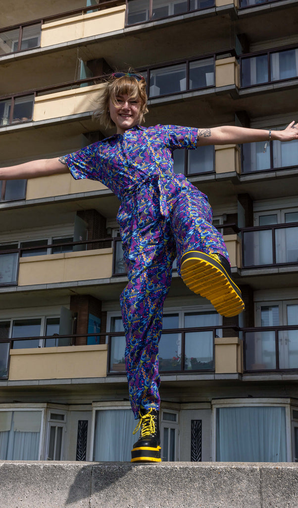 The 90's Arcade Jumpsuit being worn by a femme model with short blonde hair with koi footwear yellow and black platform boots. She is stood posing with both arms out to the side, kicking one leg forward smiling standing on top of a concrete wall in front of a block of flats. The jumpsuit print features classic 90's style shapes, squiggles and doodles in green, orange, pink, purple and light blue all on a dark blue background.