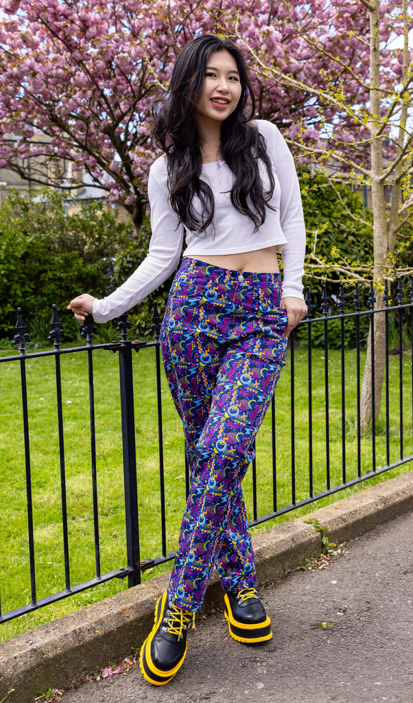 Model with long dark hair is stood outside in Hove in front of a blossom tree wearing 90's Arcade High Waist Straight Leg Jeans with a long sleeved white crop top and black and yellow boots. The jeans print features classic 90's style shapes, squiggles and doodles in green, orange, pink, purple and light blue all on a dark blue background. The model is smiling holding onto a railing with her feet crossed and one hand resting in the pocket of the jeans.