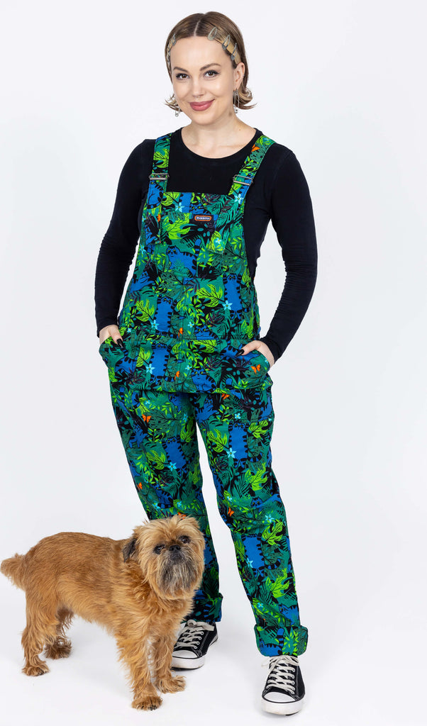 The Jungle Cats Stretch Twill Dungarees worn by a femme model with blonde bob cut hair and a black long sleeve top and black trainers on a white background. At her feet is a small brown dog looking up to camera. She is facing forward with both hands in the dungaree pockets smiling to camera. The dungaree print is a dark green base with large blue tabby cats playing amongst a jungle with various green and black foliage leaves with orange butterflies and blue flowers.