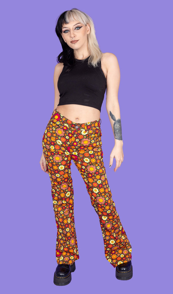 Model is wearing Ditsy Floral Bell Bottom Flares paired with a black crop top and black boots. The flares are orange and covered in red, orange and yellow flowers. The model is posing in front of a purple background facing the camera.