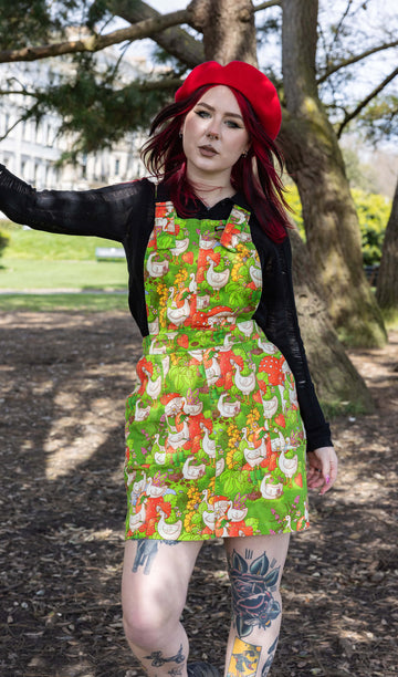 The Run & Fly x The Mushroom Babes In The Geese Garden Stretch Twill Pinafore Dress being worn by Flo outside in a park with a long sleeve black top, black trainers and red mushroom beret. She has red and black hair and tattoos, she is facing forward smising leaning one hand on a tree. The print features geese wearing various cute hats, such as mushrooms and frogs, collecting flowers and strawberries on green grass.