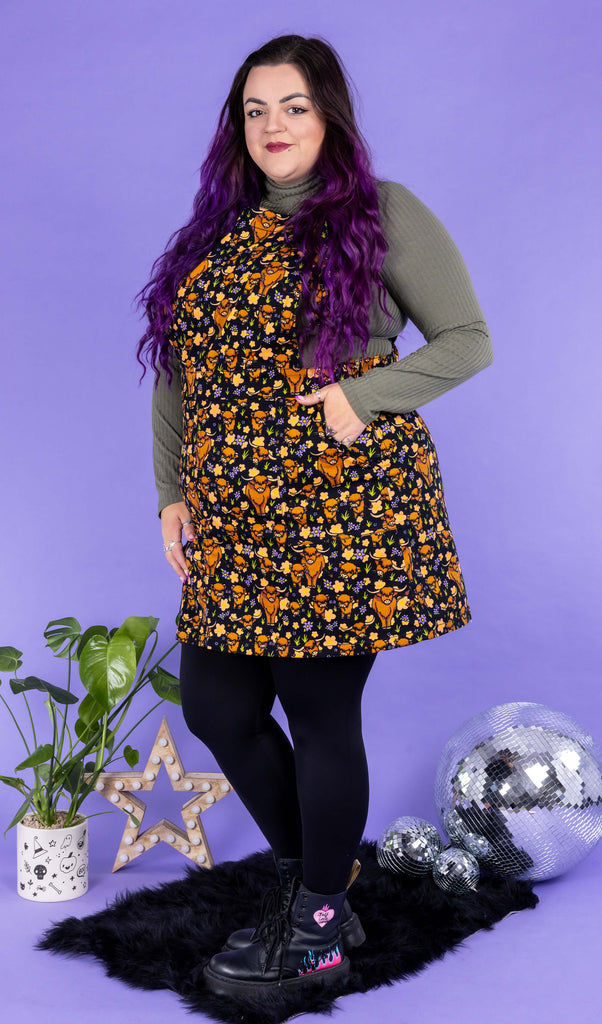 Luisa, a femme model with purple hair, is wearing Highland Cows Stretch Twill Pinafore Dress with a long sleeved khaki turtle neck underneath and black tights and boots. The black pinafore dress has an all over print of Highland cows amongst purple flowers and grass designed by Jen James. Luisa is stood on a black fluffy rug amongst disco balls, a star shaped lamp and a plant in front of a lilac backdrop and is posing to the one hand in the pinafore pocket and one leg slightly bent.
