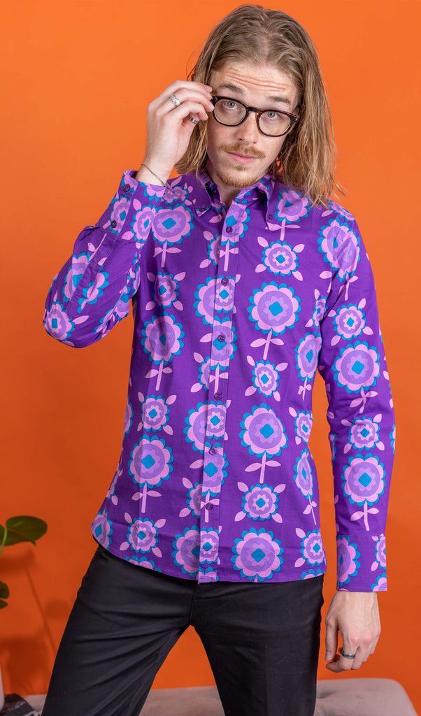 Jack, a male model with shoulder length hair and glasses is stood in front of an orange backdrop at a photography studio in Hove wearing Purple Retro Flowers Long Sleeve Shirt with black trousers. Jack is facing the camera posing with his hand on his glasses and other down by his side. The photo is cropped at the knees.
