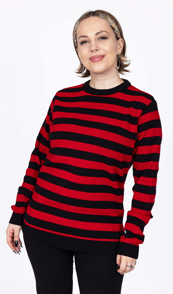 The Red and Black Striped Jumper worn by a femme model with short blonde hair with black trousers. She is facing forward smiling with her hands resting by her side. The jumper has a black neck and sleeve trim with alternating black and red stripes all over.