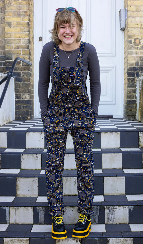 The Trash Pandas Stretch Twill Dungarees worn by a femme model with short blonde hair with a grey long sleeve top and koi footwear black and yellow platform boots. She is posing stood on a set of black and white tiled steps with both hands in the dungaree pockets smiling leaning forward. The dungaree print features cheeky illustrated racoons, trash bins, rubbish, yellow moons and stars and white sparkles on a dark blue background.