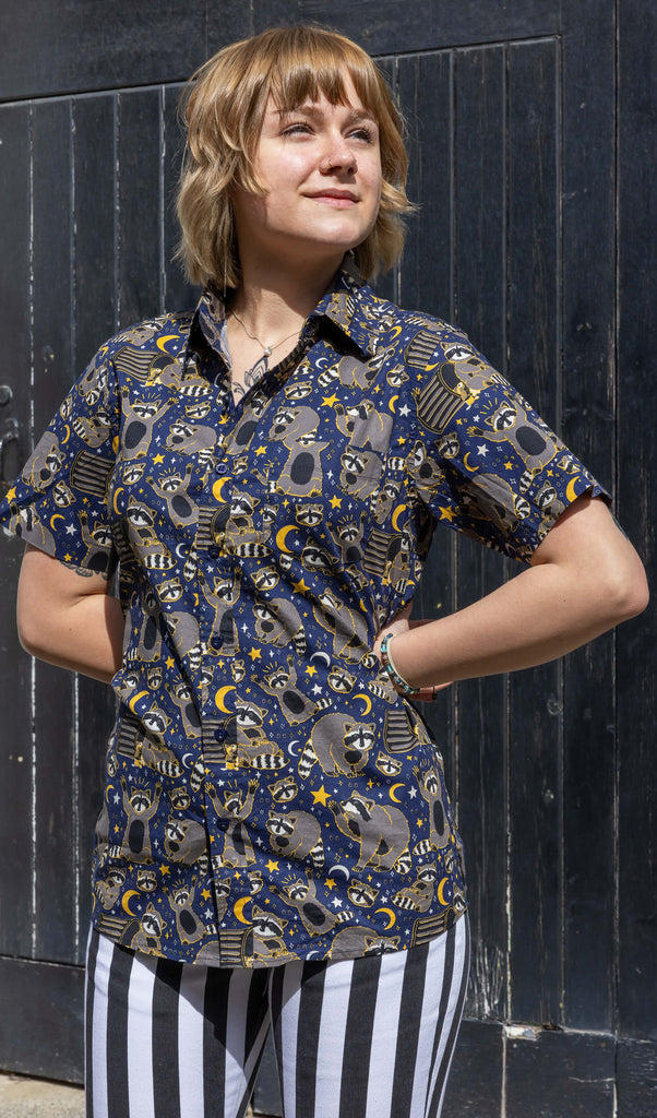 The Trash Pandas Short Sleeve Shirt worn by a femme model with short blonde hair and black and white stripe flared run and fly jeans. She is posing outside in front of a black wooden door smiling with both hands on her hips looking off to the right. The shirt print features cheeky illustrated racoons, trash bins, rubbish, yellow moons and stars and white sparkles on a dark blue background.