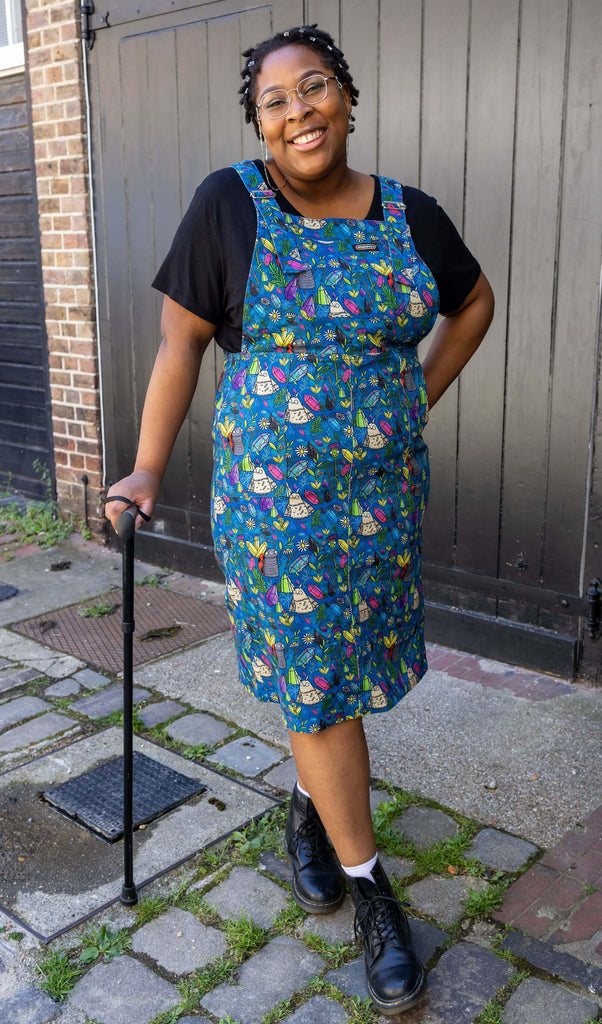 Han, a black femme model with silver glasses with a chain, is stood outside in Hove in a mews wearing Run & Fly x Katie Abey Crystal Critters Stretch Twill Long Pinafore Dress with a black t shirt underneath and black boots. The blue pinafore dress has an all over print of critters and crystals illustrated by Katie Abey. Han is smiling toward the camera with one hand in the pinafore pocket and one leg in front of the other. She is leaning on her black walking aid. 