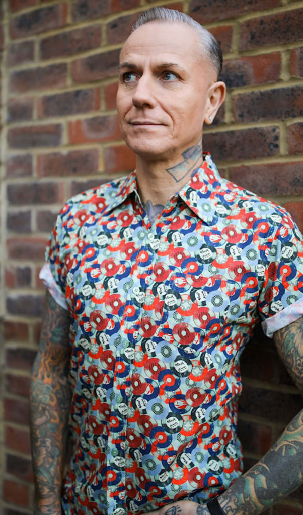 Jim is stood in front of a brick wall wearing the red and blue record short sleeve shirt with blue denim jeans. They are heavily tattooed with swept back silver hair. They are facing the camera posing with one hand on their hip whilst the other rests by their side. Photo is cropped from the hips up.