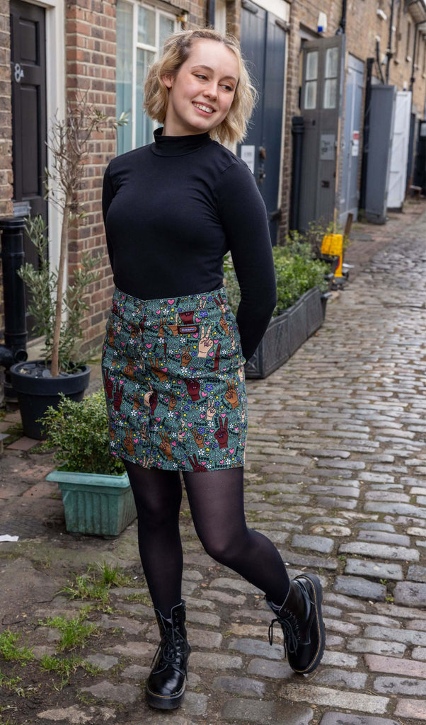 Model with short blonde hair wearing Peace and Love A Line Skirt paired with long sleeve black turtleneck top and chunky black boots. The skirt an olive green colour with hands in different skin tones making the peace sign, various flowers, peace symbols and love hearts in white, yellow, purple and pink and 'peace' and 'love' printed on. There is a Run&Fly logo on front left pocket of the skirt and it has silver buttons down the front.