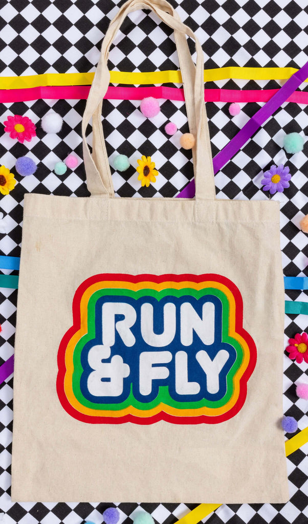 flat lay of a cream canvas tote bag with a big Run&Fly logo on the front resting on a black and white checkerboard background with colourful pom pom and flower decorations.