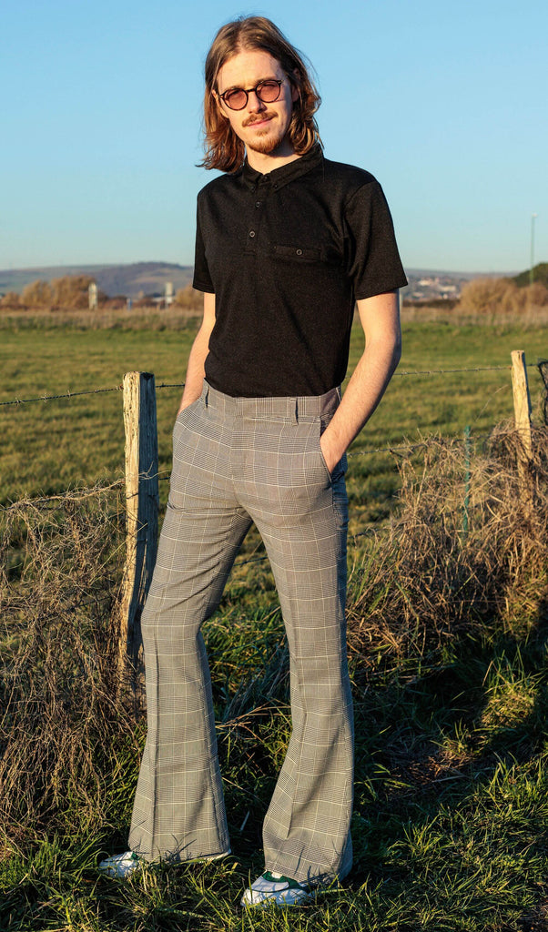 Jack is stood outside near a grass field wearing the Presley prince of Wales check bell bottom trousers with a black short sleeve shirt and white trainers. They have mid length blonde hair with circle polarised glasses. They are facing the camera posing with both hands in their front trouser pockets and one leg out to the side to highlight the flares whilst smiling.