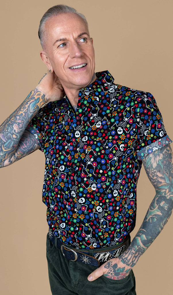 James, a white male heavily tattooed model is stood in a studio in Hove in front of a beige background wearing Halloween: Boogie Bones Short Sleeve Shirt with green corduroy trousers. Boogie bones print is a black base with multicoloured flowers and dancing smiling cartoon skeletons all over. James is posing with one hand in his pocket and other on the back of his neck.