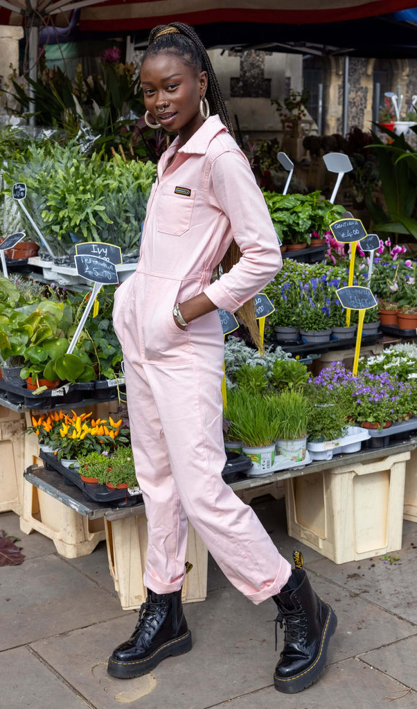 Yolande, a black femme model with piercings and a long ponytail, is stood in front of a flower stand in Hove wearing Run & Fly Pink Stretch Twill Boiler Suit with chunky black boots. Yolande is looking at the camera with one leg slightly bent and hands in the boiler suit pockets. 