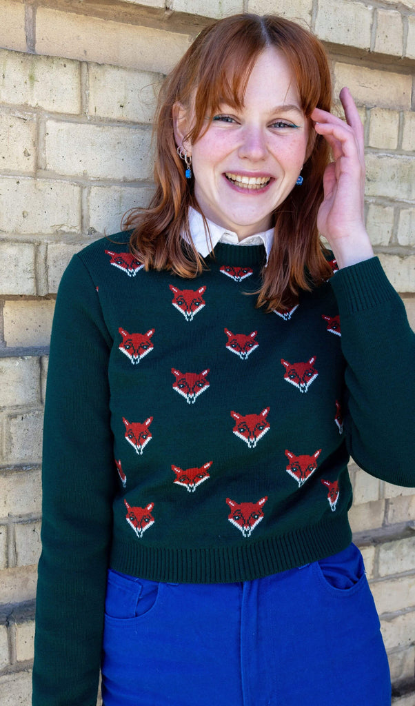 Model is wearing Green Fox Cropped Jumper, paired with a white collared shirt and bright blue jeans. The jumper is a dark forest green colour with a print of multiple red foxes on the front torso. Model is smiling at the camera with hand in her hair.