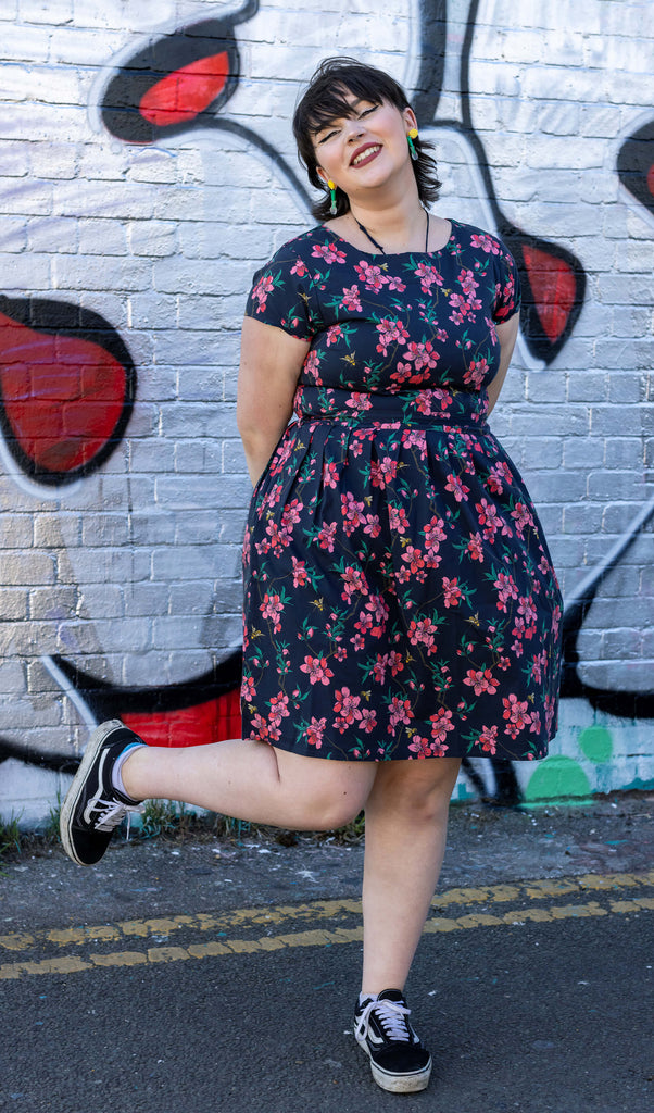 Rebecca is stood in front of a graffiti wall wearing the cherry blossom black belted stretch tea dress with pockets with black trainers. She is facing forward posing standing on one leg with her arms crossed behind her and smiling to camera.
