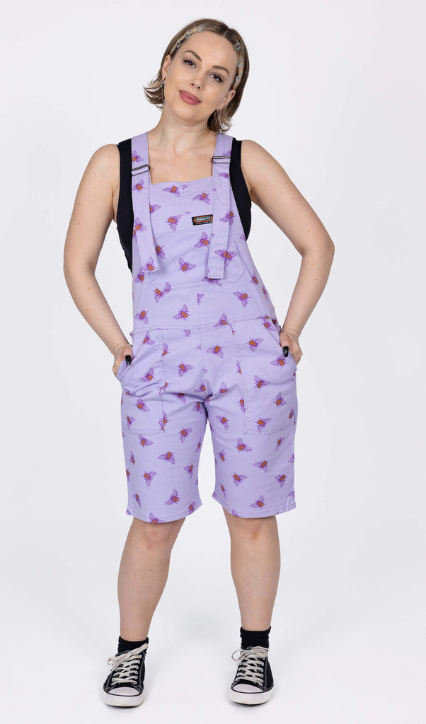 The Lavender Bees Stretch Twill Dungaree Shorts worn by a femme model with short blonde hair with a black crop top and black trainers. She is facing forward smiling with her head tilted slightly, both hands are resting on her hips in the dunga short pockets. The dungaree shorts print is a pastel purple covered with dark purple and mustard yellow bee illustrations all over.