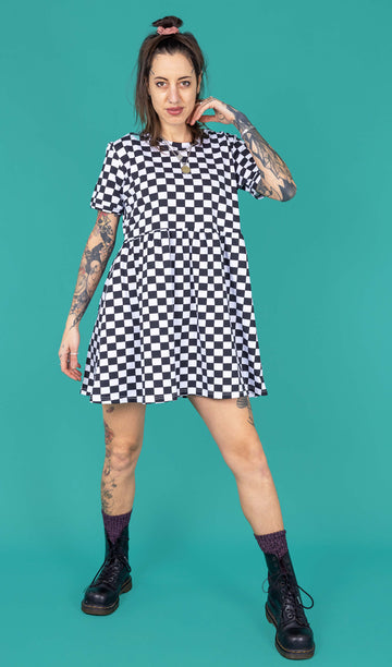 yo a white femme model with tattoos is wearing a black and white checkerboard smock dress and Doc Martens