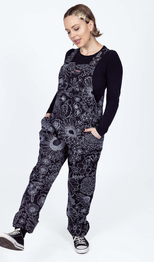 The Black and White Floral Stretch Twill Dungarees worn by a femme model with short blonde hair with a black long sleeve top and black trainers on a white background. She is facing forward with both hands in the dungaree pockets looking down kicking one leg forward. The dungaree print is a black base with various floral and botanical plants in a white outline.