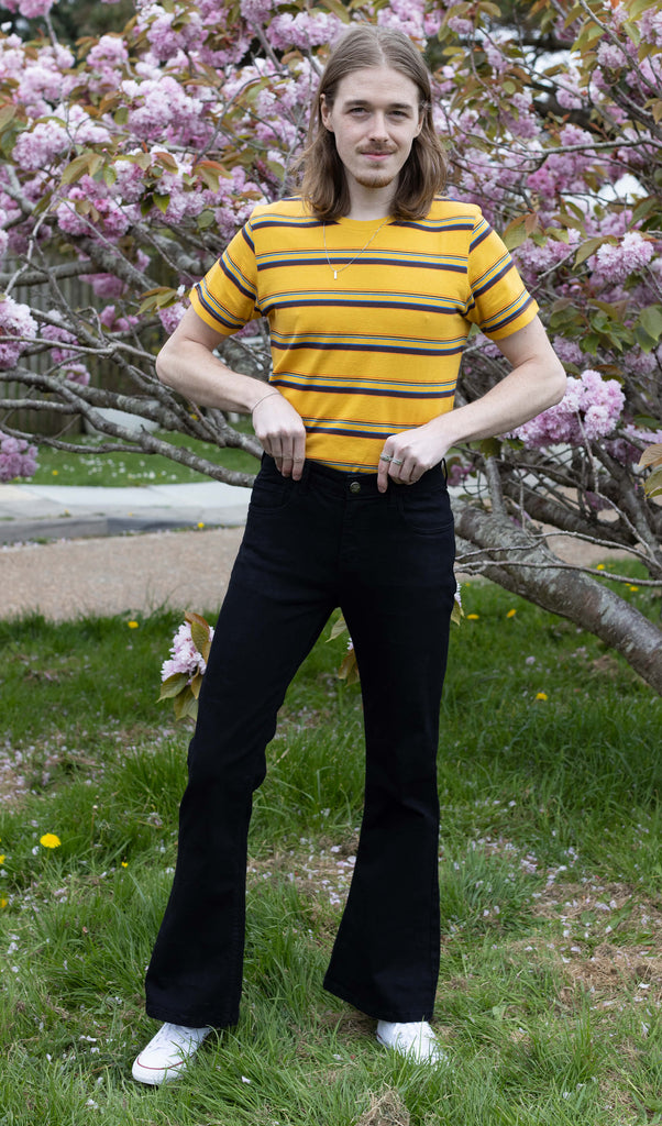 Jack is a tall blonde male with shoulder length hair looking into camera and smiling wearing a yellow retro tee and black bell bottom flares against a pink blossom tree
