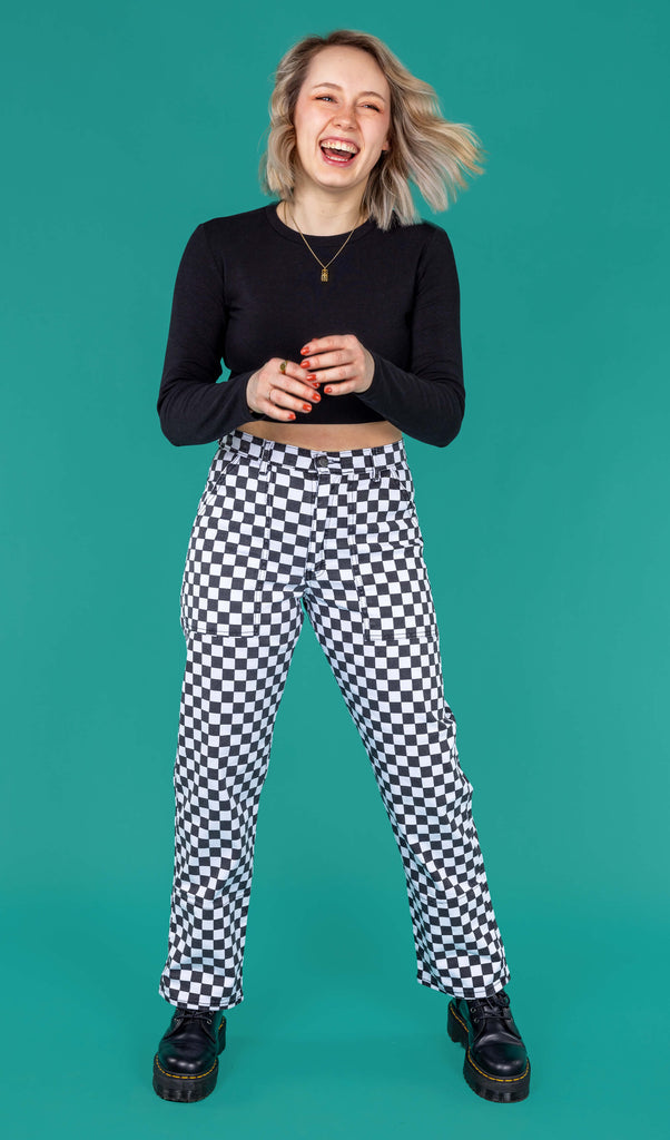 Amy a blonde white model with short hair is smiling and laughing wearing a black long sleeved top and black and white checkerboard trousers with black boots on a green background