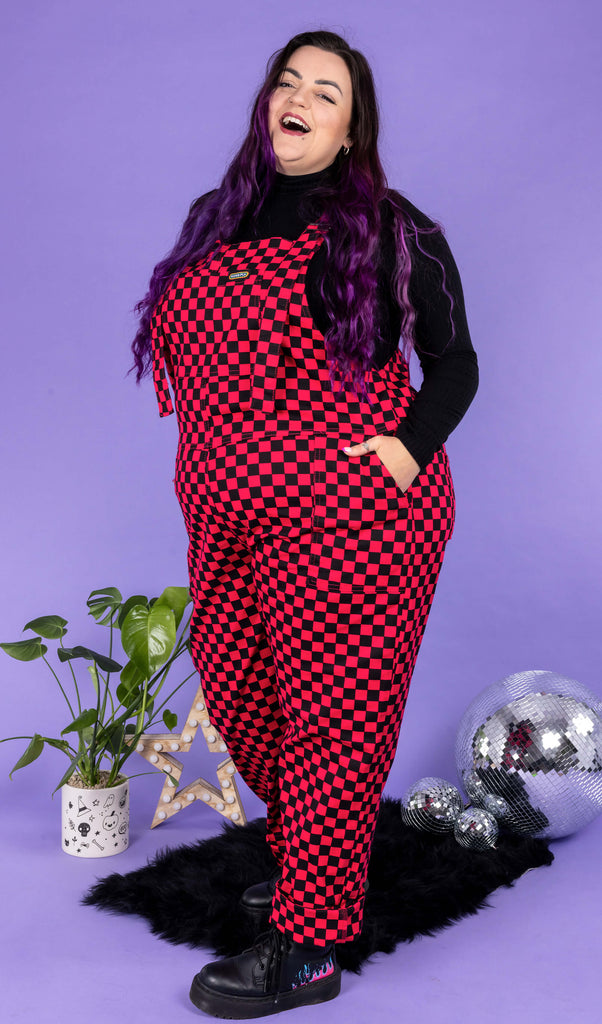 Luisa, a white femme model with purple hair, is wearing Black & Viva Magenta Checkerboard Stretch Twill Dungarees with a long sleeve black turtle neck underneath and black boots. Luisa is stood in front of a lilac background on a black fluffy rug amongst disco balls, a star shaped light and a plant and is facing the camera laughing with her hands in the dungaree pockets.