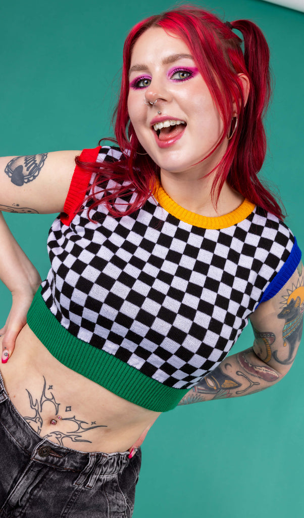 Flo wearing the Run and Fly Colour Block Checkerboard Cropped Knitted Tank Top on a teal background. She has styled the all over black and white checkerboard vest with a yellow trim neck, red left arm trim, blue right arm trim and green hem trim with oversized y2k cargo trousers, silver hoop earrings and neon pink eye makeup. Flo has red hair in bunches with piercings and tattoos. She is facing forward leaning in smiling with both hands on her hips.