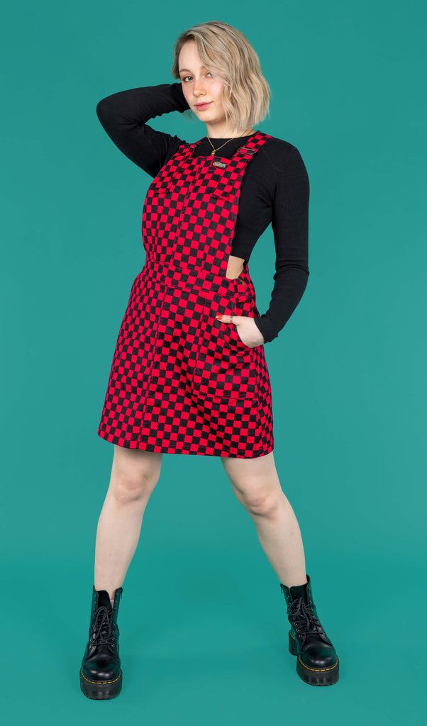 Amy a blonde white femme model with shoulder length hair is wearing a black long sleeved top and black boots with a pink and black pinafore dress stood against a green background