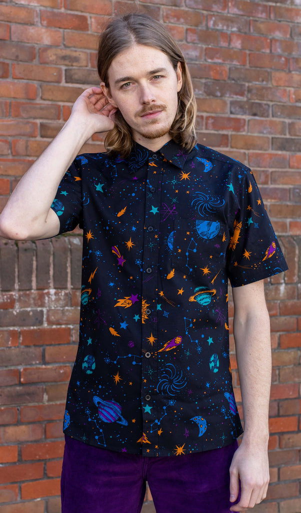 Jack is stood in front of a brick wall wearing the cosmic space short sleeve shirt with purple trousers. They are facing forward posing with one hand tucking their hair behind their ear whilst the other hand rests by their side. Photo is cropped from the hips up.