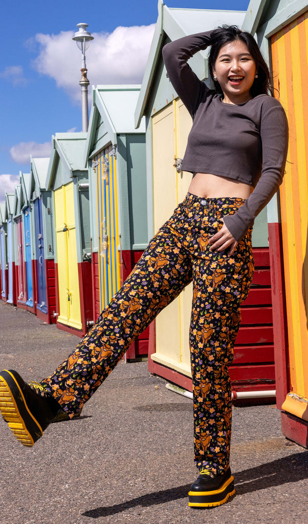 The Highland Cow High Waist Straight Leg Jeans on a femme model with long black hair with yellow and black platform boots and long sleeve grey top in front of colourful beach huts. The straight leg trousers are a black base covered in brown highland cows, tan flowers, purple flowers and grass. She is leaning back on one leg laughing with one hand in her hair.