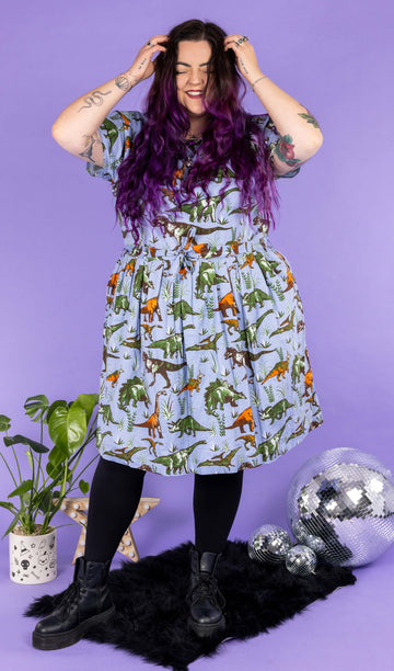 Luisa, a femme model with purple hair and tattoos, is stood in a photography studio in Hove in front of a purple backdrop wearing Jurassic Adventure Dinosaur Tea Dress With Pockets with black tights and boots. The light blue dress has an all over dinosaur print with a matching fabric tie belt. Luisa is stood on a black furry rug amongst disco balls, a star light and a plant and is facing the camera smiling with her hands in her hair.