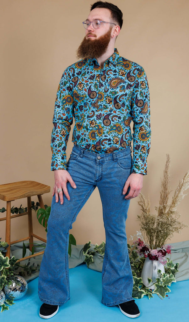 Dec a white male in his 20s with a beard and glasses is stood in a photography studio in Hove wearing New Fit Retro Stretch Stone Wash Denim Hendrix Paisley Bell Bottom Flares with Sky Blue Paisley Short Sleeve Shirt and black shoes. Dec is posing facing the camera with one leg bend and is looking off into the distance.