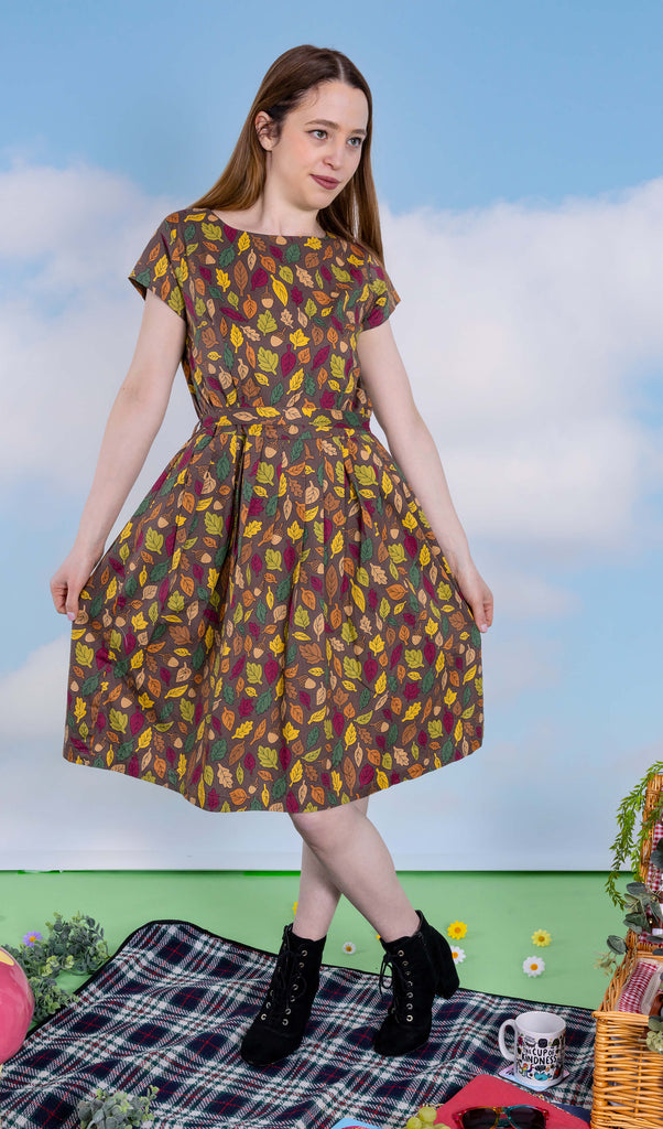 The Autumn Leaves Stretch Belted Tea Dress with Pockets on a femme model with long brown hair and black heeled boots. She is posing with one leg crossed over holding out the skirt smiling looking right at a picnic setup with a picnic basket, fruit, flowers and katie abey mugs. The dress print is a brown base with green, red, orange and yellow autumnal leaves and acorns.