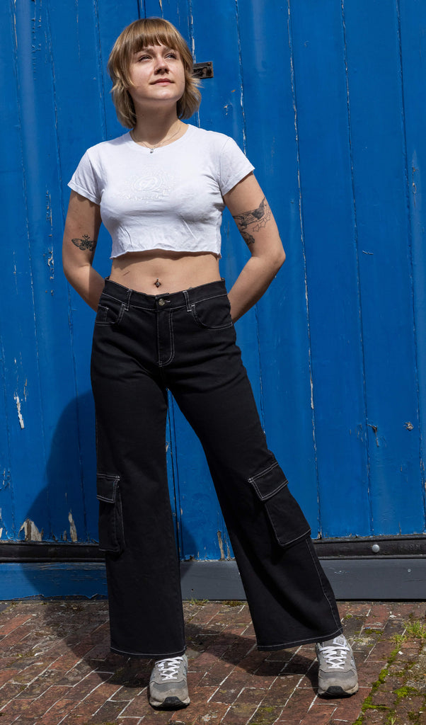 The Black Contrast Stitch Wide Leg Stretch Cargo Trousers modelled by a femme person with short blonde hair, a white crop top baby tee and grey trainers. She is stood posing in front of a blue wooden door outside leaning on her left leg, both hands resting behind her hips smiling looking off to the right. The black cargo trousers have white contrast stitching with two front flap pockets on the lower legs.