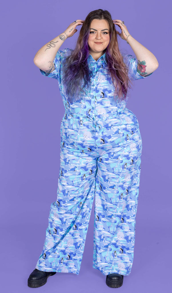 a model with purple hair wearing Winter Friends Stretch Jumpsuit paired with black boots. The jumpsuit is a light blue colour with ice burgs, seals, polar bears and penguins on. The model is posing with her hands in her hair and popping her hip to the side. The background of the photo is purple.