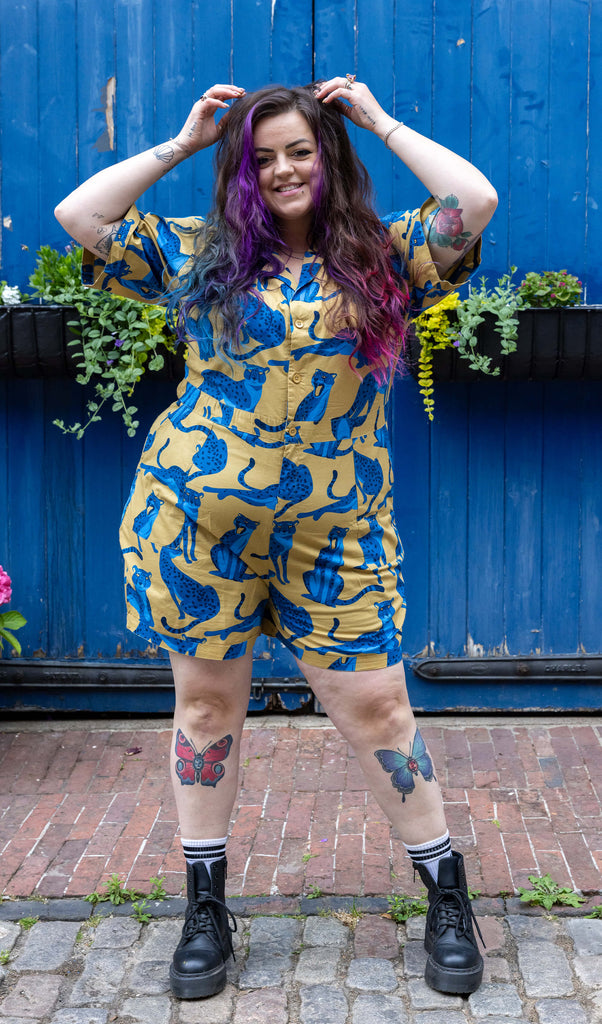Tattooed model with colourful hair wearing Big Cats Stretch Playsuit paired with chunky black boots. The playsuit is a mustard yellow colour with big blue cats printed on it in various poses. The playsuit also has buttons down the front. The model is posing and running her hands through her hair.
