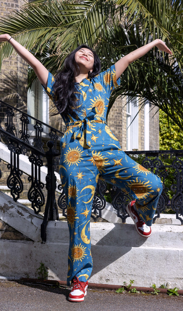 Model is stood outside in Hove in the sun in front of a town house wearing Celestial Sun and Moon Jumpsuit with red and white trainers. The blue base shirt features retro style gold moons and suns with faces, yellow sparkles and stars all over. The model is smiling with her arms in the air and one leg bent.