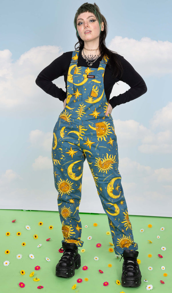 Faeryn is stood in a photography studio in Hove on a green floor with flowers scattered around. wearing Celestial Sun and Moon Stretch Twill Dungarees with a long sleeved black top underneath and chunky black shoes. The dungaree print is a denim blue colour with gold and yellow retro style suns and moons with faces, shooting stars, stars and sparkles.They are posing at the camera with their hands on their hips and feet slightly apart. The background of the photo is a blue sky.