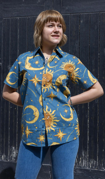 The Celestial Sun and Moon Short Sleeve Shirt worn by a femme model with short blonde hair and blue denim flare jeans from run and fly. She is posing in front of a black wooden door outside smiling with both hands on her hips looking off to the right. The blue base shirt features retro style gold moons and suns with faces, yellow sparkles and stars all over. 