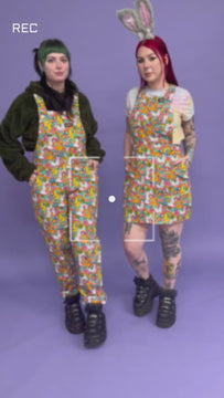 flo and Faeryn both tattooed white models wearing matching Waffle Mama Design print dungarees and a pinafore in multi colours. Faeryn is holding a carrot and squinting