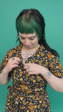 Faeryn, a tattooed model with green and black hair is wearing a black jumpsuit with various sizes of brown highland cows on it, some eating grass. There is also various peach, purple and yellow flowers on the design. The jumpsuit is paired with black boots. Faeryn is zipping up the jumpsuit to the top and twirling with their hands in the jumpsuit pockets and then flips their hair. The background of the video is green. 