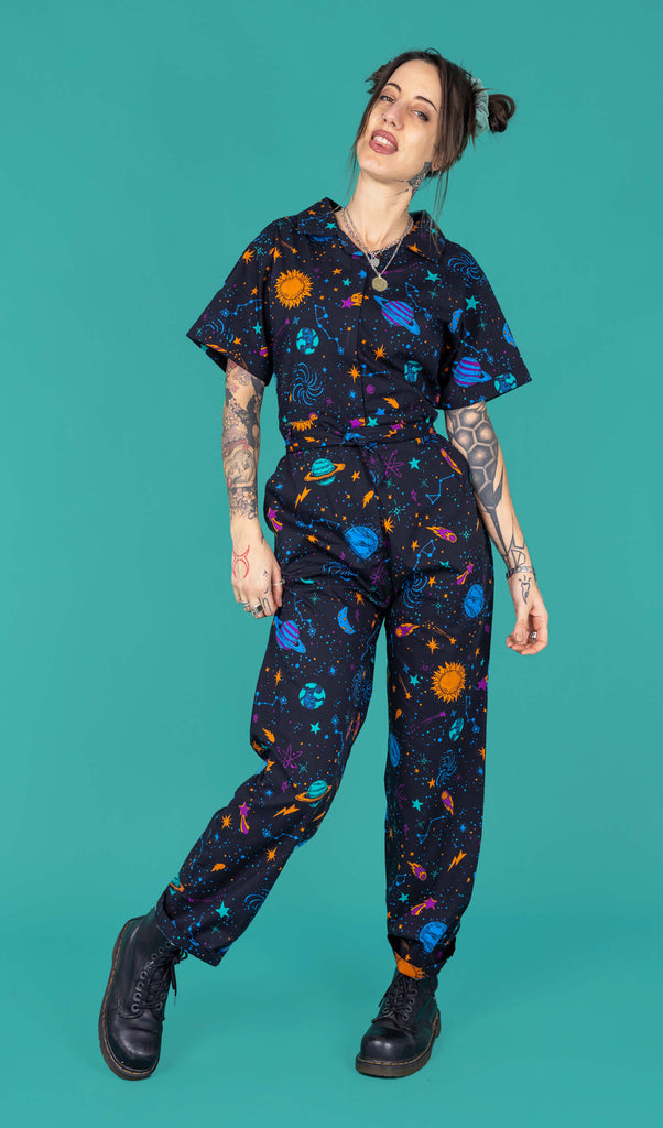 Yo a femme person with hairpin bunches and tattoos is wearing a navy jumpsuit with space print on it with short sleeves. It is shot in a studio against a green background