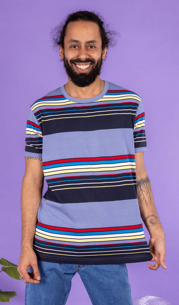 Richard, a hispanic male model with dark hair in a bun and a beard, is stood in a photography studio in Hove in front of a lilac backdrop wearing Vintage Blue Stripe Tee with blue jeans. The blue t shirt has horizontal lines in red, blue, white, yellow and navy. Richard is facing the camera smiling and is holding the bottom of the t shirt with one hand and other resting down by his side. The photo is cropped at the thighs.