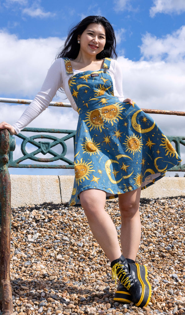 The Celestial Sun and Moon Flared Pinafore Dress worn by a femme model with long black hair with yellow and black platform boots and a white tshirt underneath. She is posing leaning on a metal railing smiling to camera on a pebble beach with a bright blue sunny cloudy sky behind. The pinafore print is a denim blue colour with gold and yellow retro style suns and moons with faces, shooting stars, stars and sparkles.