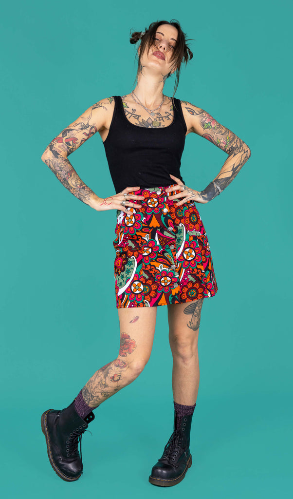 Yo a tattooed femme model is wearing a black vest and swirly floral A line skirt in a studio against a green background