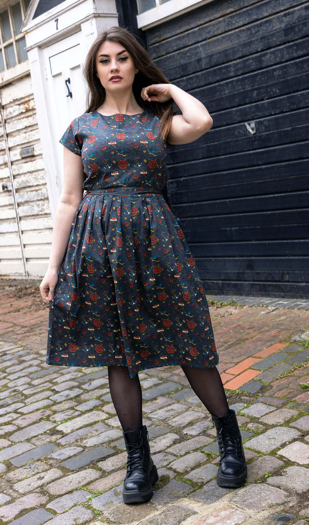 Charlotte is wearing the Run & Fly Retro Tattoo Stretch Belted Tea dress with pockets, grey with red roses, hearts and birds print, and waist belt. Model is looking at the camera, and standing in a cobbled street in front of garage doors with one hand in her hair.