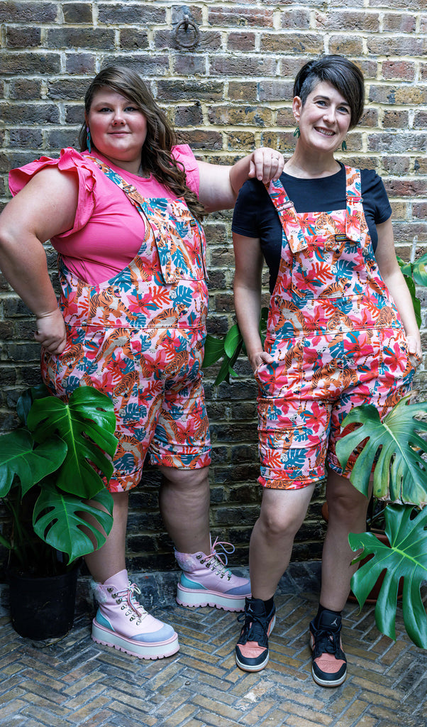Two femme models wearing the tiger lily stretch twill dungaree shorts outside amongst plants. They are both smiling and posing towards the camera.