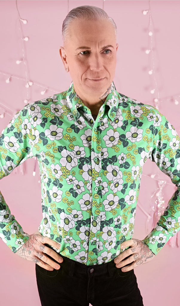 Jim is stood in front of a pink studio background wearing the peppermint poppy button down long sleeve shirt with black trousers. They are facing the camera posing with both hands on their hips, they're looking off to the right smiling. Photo is cropped from the hips up.