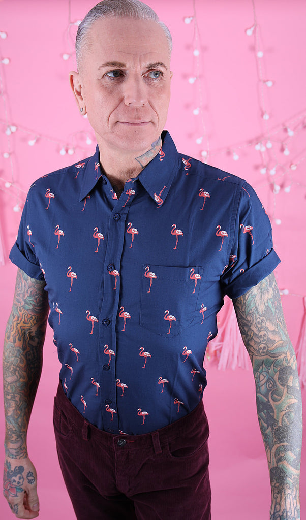 Jim is stood in front of a pink studio background wearing the preppy flamingo short sleeve shirt with burgundy trousers. They are heavily tattooed and have swept back silver hair. They are facing the camera posing with both arms out by their side and looking off to the right. Photo is cropped from the hips up.